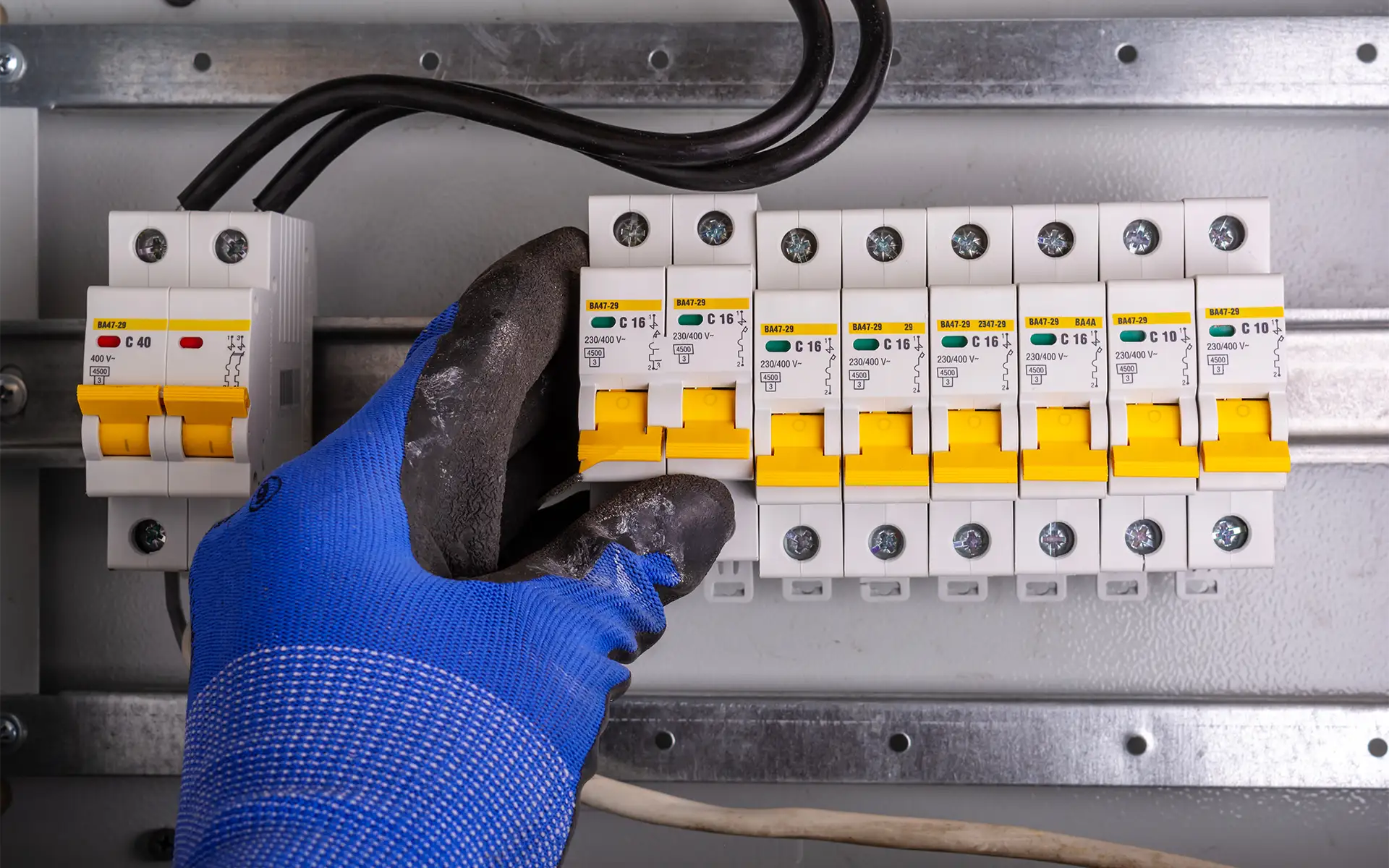 Close-up view of an electrician's gloved hand checking circuit breaker connections in a distribution box with multiple yellow switches